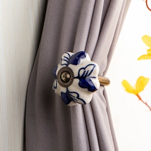 Könnte beinhalten: Ein weißer und blauer Keramik-Vorhang-Halter in Blumenform mit einem goldenen Metallring. Der Halter hält einen grauen Vorhang.