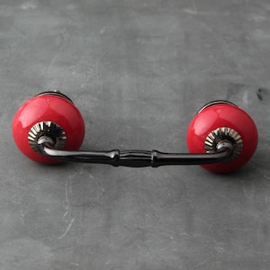 May include: Red ceramic drawer pull with a black metal bar connecting the two knobs.