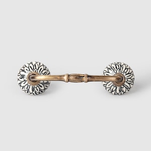 May include: A white ceramic drawer pull with a black floral design. The pull has a gold-toned metal bar connecting the two ceramic knobs.