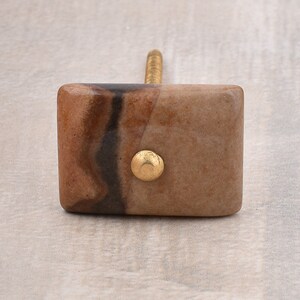 May include: A square-shaped cabinet knob with a brown and black marbled design. The knob has a gold metal accent in the center.