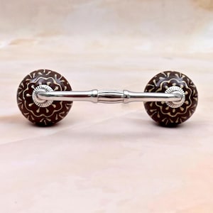 May include: A drawer pull featuring two round, dark brown ceramic knobs with a cream-colored floral pattern. The knobs are connected by a horizontal, polished silver-toned metal bar with a central decorative detail and fluted silver accents.
