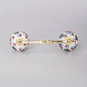 May include: A white ceramic cabinet knob with a gold metal bar connecting two round knobs. The knobs are decorated with a blue, red and green floral pattern.