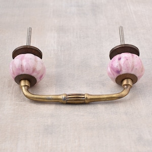 以下が含まれることがあります： 真鍮製のハンドルと2つのピンク色のセラミックノブが付いたキャビネットプル。ノブは紫色のアクセントが付いた斑点模様です。プルは、ねじ付きロッドで簡単に取り付けられるように設計されています。