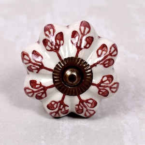 May include: A white ceramic drawer knob with a scalloped, flower-like shape. The surface features a hand-painted reddish-brown botanical pattern of stems and leaves radiating from the centre. A circular, ribbed antique brass metal fitting is centred.