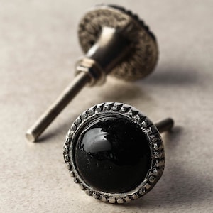 Könnte beinhalten: Zwei silberne Schrankknöpfe mit einem dekorativen Rand. Ein Knopf hat eine schwarze Mitte und der andere eine silberne Mitte.