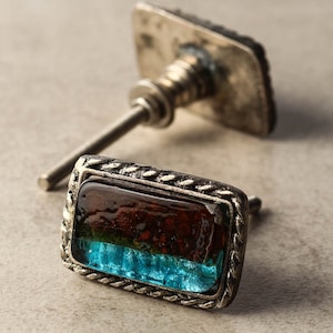 Puede incluir: Pomos decorativos para armarios con un inserto de vidrio rectangular multicolor. El vidrio presenta tonos de marrón, verde y azul. Los pomos tienen un marco de metal plateado y una placa trasera cuadrada. Herrajes para muebles.