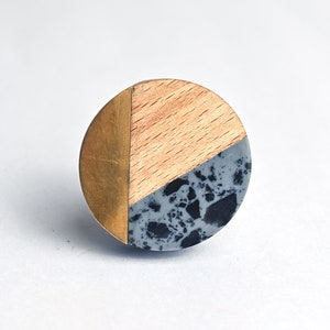 Könnte beinhalten: Ein runder Schrankknopf mit einem Design aus gemischten Materialien. Der Knopf hat einen Abschnitt aus hellem Holz, einen Abschnitt aus Messing und einen Abschnitt aus grau gesprenkeltem Material. Der Knopf ist für Möbel und Heimdekoration konzipiert.