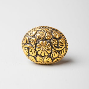 May include: A gold-coloured button with a detailed floral design. The button has a raised, textured surface and a black outline.