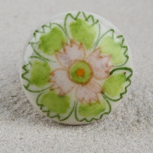 Può includere: Pomello per armadio rotondo in ceramica bianca con un motivo floreale dipinto a mano. Il fiore presenta un centro rosa, foglie verdi e un centro arancione. Elemento decorativo per mobili.