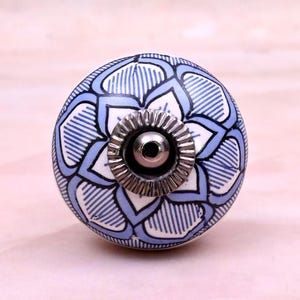 Op de afbeelding: Een ronde keramische meubelknop met een witte basis en een blauw bloemmotief van gelaagde bloemblaadjes. Het midden van de knop heeft een metalen zilverkleurig rond onderdeel met een geribbelde rand en een klein centraal gat.
