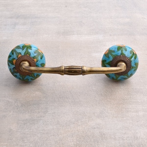 May include: A decorative cabinet pull with blue, green, and orange floral patterned ceramic knobs. The knobs are connected by a brass-colored handle. The pull is designed for use on drawers or cabinet doors.