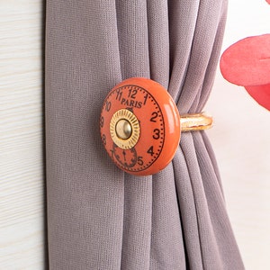 Könnte beinhalten: Orangefarbener Vorhang-Halter aus Keramik mit einem goldenen Metallring. Der Halter hat ein Zifferblatt mit den Worten "Paris" und den Zahlen 1 bis 12.