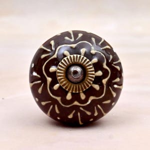 Op de afbeelding: Een ronde keramische meubelknop met een donkerbruine glanzende afwerking en een crèmekleurig bloemmotief. Het midden van de knop is voorzien van een cirkelvormige messingkleurige metalen plaat en een kleine, verhoogde zilverkleurige boutkop.