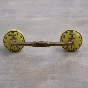 以下が含まれることがあります： 金色の金属棒で繋がれた、黄色のセラミック製の引き出しノブ2個。それぞれのノブには、"Paris Clocks"という文字と黒いローマ数字の目盛りが付いた時計の文字盤があります。