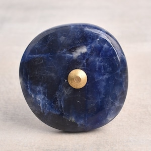 Op de afbeelding: Een ronde, blauwe en gouden knop gemaakt van natuursteen. De knop heeft een glad, gepolijst oppervlak en een gouden metalen accent in het midden.