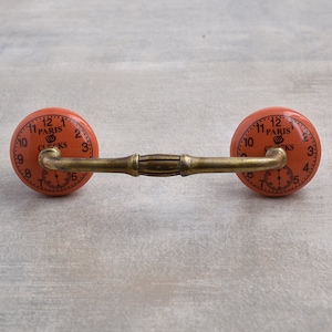 以下が含まれることがあります： 金色の金属製のつなぎ棒が付いたオレンジ色のセラミック製の引き出しノブ2個。各ノブには、「Paris Clocks」という文字とローマ数字の目盛りが付いた時計の文字盤があります。