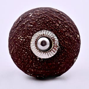 Puede incluir: Un pomo de mueble redondo con una superficie texturizada de color marrón oscuro que recuerda a la corteza natural. El centro presenta una placa metálica circular plateada con un patrón estriado que rodea un núcleo metálico pulido.