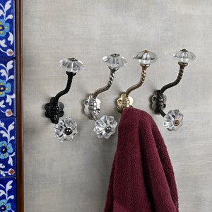 Może przedstawiać: Cztery ozdobne haczyki z kryształowymi gałkami w kolorze czarnym, srebrnym, złotym i mosiężnym. Haczyki są zamontowane na szarej ścianie, a na jednym z nich wisi bordowy ręcznik.