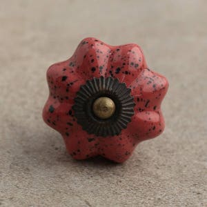 Op de afbeelding: Een rode keramische knop met zwarte stippen en een stervormig ontwerp. De knop heeft een messing centrum en een zwarte metalen ring eromheen.