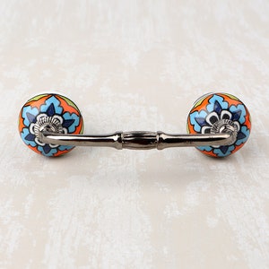 May include: A decorative cabinet pull with two round ceramic knobs. The knobs feature a floral design in blue, orange, and white against a black background. The connecting bar is a dark, metallic color.