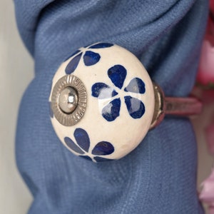 Könnte beinhalten: Weißer Keramik-Knopf mit blauem Blumenmuster und einem silbernen Metallring. Der Knopf befindet sich auf einem blauen Stoffhintergrund.