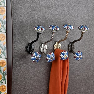 May include: Five decorative hooks with a black metal base and a blue and white ceramic knob. The hooks are mounted on a gray wall and a single orange scarf is hanging from one of the hooks.