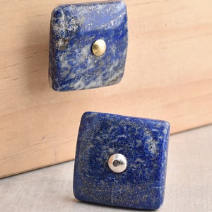 Może przedstawiać: Dwa kwadratowe gałki do szafek wykonane z niebieskiego kamienia lapis lazuli z złotymi i srebrnymi akcentami.