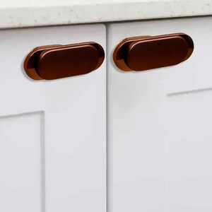 May include: Two bronze-colored, oval cabinet pulls mounted on a white cabinet door. The pulls have a smooth, metallic finish and a slightly raised profile. The cabinet doors are part of a kitchen or bathroom setup.