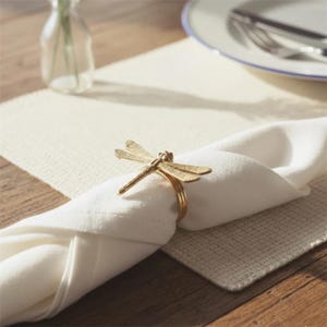 Könnte beinhalten: Ein goldfarbener Serviettenring in Form einer Libelle schmückt eine gefaltete weiße Stoffserviette. Die Serviette liegt auf einem cremefarbenen Platzset, im Hintergrund sind ein Teller und Besteck zu sehen. Die Libelle hat detaillierte Flügel und einen Ringfuß.