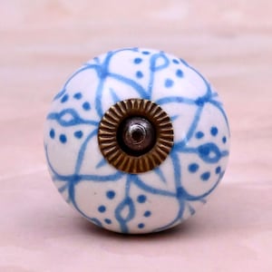 Op de afbeelding: Een ronde keramische meubelknop met een witte basis en een handgeschilderd blauw bloemen- en stippenpatroon. Het midden van de knop heeft een cirkelvormig, geribbeld metalen beslag in antiek messing met een donkere schroefkop in het midden.