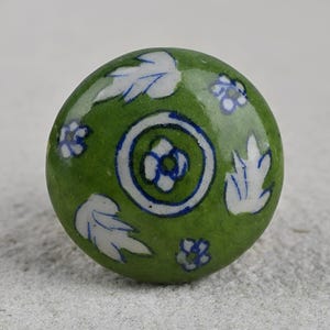 May include: Green ceramic knob with a white and blue floral design. The knob has a circular shape and is decorated with stylized leaves and flowers.