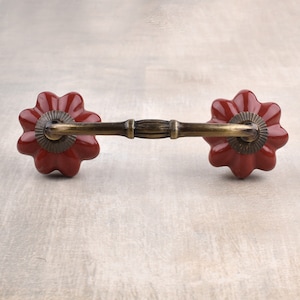 May include: A red ceramic drawer pull with a brass bar connecting two flower-shaped knobs.