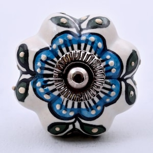 May include: A flower-shaped ceramic cabinet knob with a white base, featuring hand-painted blue petals, dark green leaves, and small white dots. A circular, ridged silver-tone metal hardware piece is centered on the front of the knob.