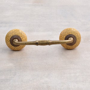 May include: A pair of ceramic drawer pulls with a crackled glaze finish in a light yellow color. The pulls have a brown accent ring and are attached to a brass bar.