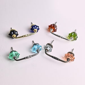 Könnte beinhalten: Sortiment von sechs Schrankknöpfen mit verschiedenen farbigen Glasakzenten. Die Knöpfe haben eine Messing- oder schwarze Metallbasis und einen gebogenen Griff.