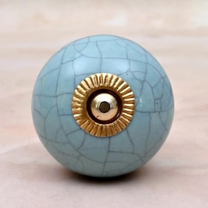 May include: A round cabinet knob with a light blue crackle glaze finish. The center features a circular gold-colored metal plate with a ridged, sunburst-like texture surrounding a smooth, raised gold-colored metal dome in the middle.