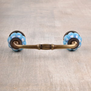 May include: A drawer pull featuring a curved, antique brass-finished metal handle connected to two round, fluted ceramic knobs. The knobs are white with a light blue, cloud-like pattern and are mounted on circular brass-toned metal bases.
