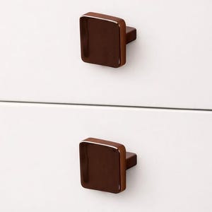 May include: A white cabinet featuring two dark brown, square cabinet pulls with a glossy finish. The pulls are mounted on the cabinet doors. The cabinet top is a light wood colour. The pulls are approximately 2.5 cm square.