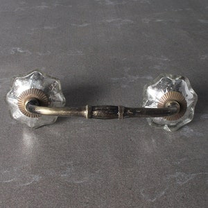 May include: A silver-toned metal drawer pull with a curved design and two decorative glass knobs. The knobs have a scalloped edge and a raised center.