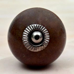 May include: A spherical brown knob with a smooth, mottled surface texture. The center features a circular, silver-toned metal plate with a ridged, sunburst-like pattern surrounding a small, raised, polished silver-toned dome in the middle.