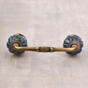 May include: A brass cabinet pull with two blue and green ceramic knobs. The knobs have a floral pattern and are attached to a curved brass bar.
