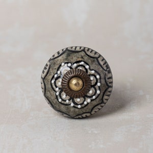 Puede incluir: Un pomo de cerámica gris con un diseño floral en blanco y negro y un centro dorado. El pomo tiene un borde ligeramente elevado.