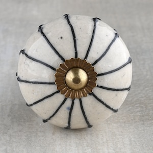 Op de afbeelding: Witte keramische knop met zwarte lijnen die uitstralen van een gouden middelpunt. De knop heeft een getextureerd oppervlak.