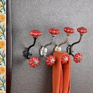 Puede incluir: Cinco ganchos de cerámica roja en forma de flor con una base de metal dorado y negro montada en una pared gris. Una larga bufanda naranja cuelga de uno de los ganchos.