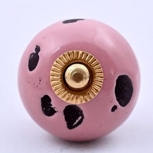 Può includere: Un pomello sferico per mobili con una finitura rosa liscia e lucida e macchie nere irregolari sparse sulla superficie. Il centro presenta un componente in metallo dorato, circolare, con un bordo scanalato e un perno centrale.