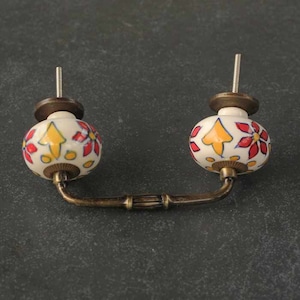 May include: A pair of ceramic drawer pulls with a brass connecting bar. The pulls are round and feature a white background with red and yellow floral designs.