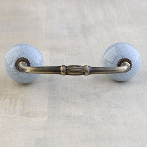 May include: A light blue ceramic drawer pull with a distressed finish and a brass bar connecting the two knobs.