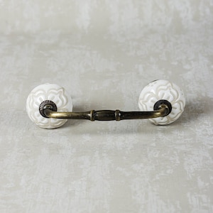 May include: White ceramic drawer pull with a floral design and a brass bar connecting the two knobs.