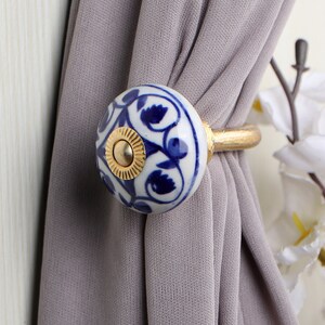 May include: A white ceramic curtain tieback with a blue and gold floral design. The tieback is attached to a gold metal ring.