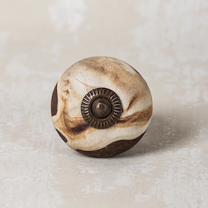 May include: A round, brown and white ceramic knob with a brass accent. The knob has a swirling pattern.